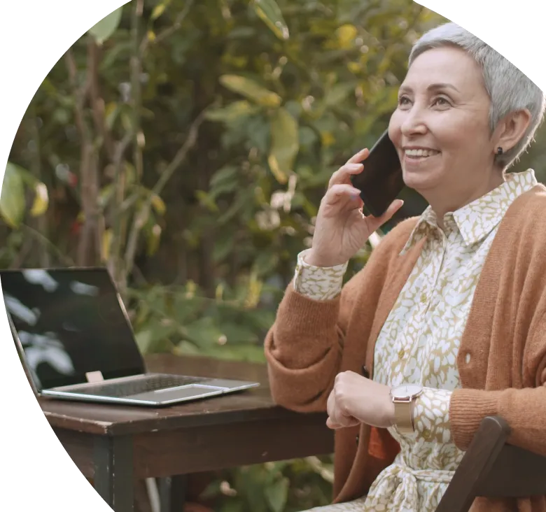 A smiling woman talking on the phone at a garden table with her laptop open